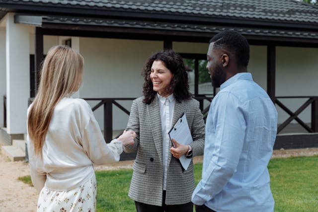 Two people shaking hands outside while one holds a clipboard and a third person stands near them